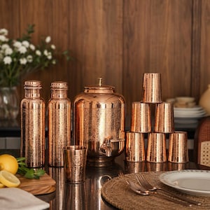 May include: A copper-colored drinkware set, including bottles, a water dispenser with a spigot, and tumblers. The set has a textured surface and is arranged on a dark surface. A wooden cutting board with lemons and a vase of white flowers are also present.