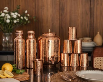 Puede incluir: Un juego de recipientes para bebidas de color cobre, que incluye botellas, un dispensador de agua con grifo y vasos. El juego tiene una superficie texturizada y está dispuesto sobre una superficie oscura. También hay una tabla de cortar de madera con limones y un jarrón con flores blancas.