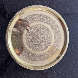 Puede incluir: Una bandeja redonda de metal dorado con un intrincado dise&ntilde;o floral grabado en la superficie. La bandeja tiene un borde con cuentas y un acabado pulido, adecuado para servir o exhibir.