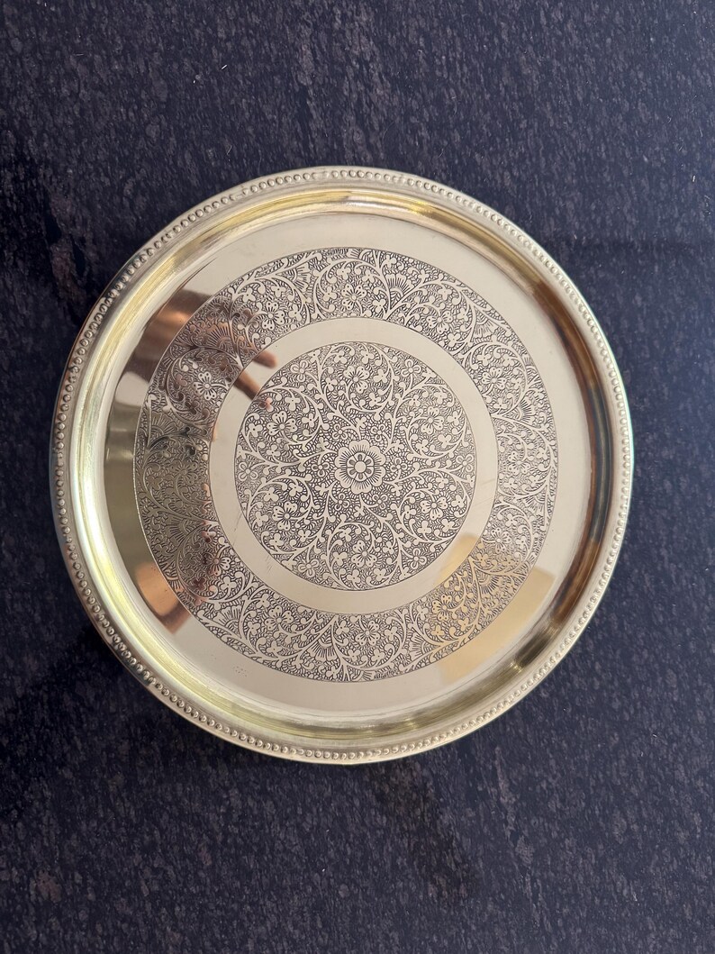Puede incluir: Una bandeja redonda de metal dorado con un intrincado dise&ntilde;o floral grabado en la superficie. La bandeja tiene un borde con cuentas y un acabado pulido, adecuado para servir o exhibir.