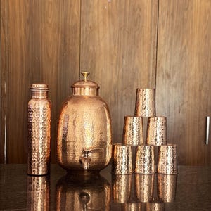 May include: A copper water set with a hammered finish. The set comprises a tall bottle, a large water dispenser with a tap, and six matching cups. The items are arranged on a reflective surface, set against a wooden background.