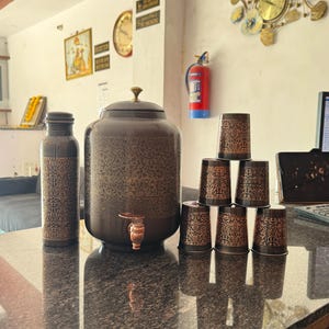 May include: A dark brown water dispenser set with a matching bottle and six cups. The dispenser has a spigot and is adorned with an intricate floral pattern. The cups are stacked, and the set is on a reflective surface.