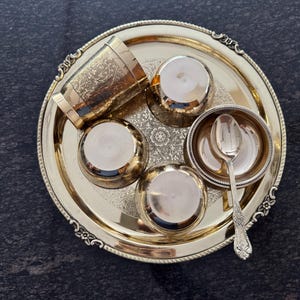 Puede incluir: Un juego de servir de metal dorado con una bandeja, una taza, cuencos peque&ntilde;os y una cuchara. La bandeja y la taza tienen dise&ntilde;os florales ornamentados. El conjunto est&aacute; dispuesto sobre una superficie oscura y texturizada.
