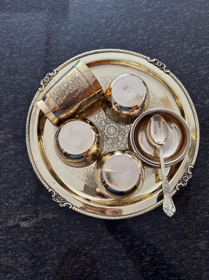 Puede incluir: Un juego de servir de metal dorado con una bandeja, una taza, cuencos peque&ntilde;os y una cuchara. La bandeja y la taza tienen dise&ntilde;os florales ornamentados. El conjunto est&aacute; dispuesto sobre una superficie oscura y texturizada.