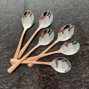 May include: A set of six spoons with polished silver bowls and copper-colored, textured handles. The spoons are arranged in a fan shape, reflecting the surrounding environment. The spoons are designed for serving or eating.