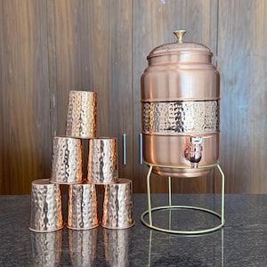 Puede incluir: Un dispensador de agua de cobre con grifo y tapa, sobre un soporte dorado. Seis tazas de cobre a juego con una textura martillada están apiladas cerca. El dispensador y las tazas tienen un acabado metálico cálido.