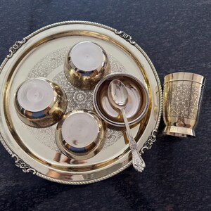 Puede incluir: Un juego de servir de metal dorado, que incluye una bandeja redonda con bordes ornamentados, cuatro cuencos peque&ntilde;os, una cuchara y una taza decorativa. La bandeja y la taza presentan patrones florales grabados. El conjunto est&aacute; dispuesto sobre una superficie oscura.