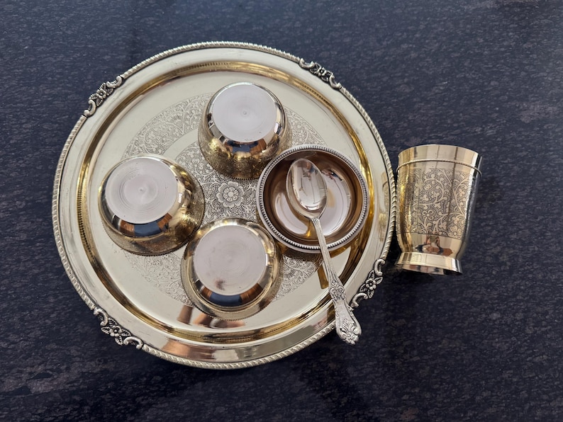 Puede incluir: Un juego de servir de metal dorado, que incluye una bandeja redonda con bordes ornamentados, cuatro cuencos peque&ntilde;os, una cuchara y una taza decorativa. La bandeja y la taza presentan patrones florales grabados. El conjunto est&aacute; dispuesto sobre una superficie oscura.