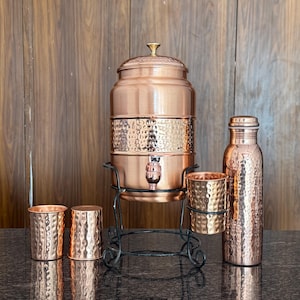 May include: A copper water dispenser with a tap, a matching bottle, and three copper cups. The dispenser sits on a black metal stand. The copper items have a hammered texture and are displayed on a dark surface.