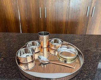 Puede incluir: Un juego de thali de metal martillado en tono cobre, con una bandeja redonda grande, cuencos pequeños, una taza alta, una cuchara y un anillo decorativo. El juego está dispuesto sobre una superficie oscura y moteada.
