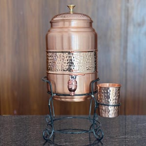 May include: A copper water dispenser with a hammered texture, a tap, and a lid, set on a black wrought iron stand. A matching copper cup sits beside it. The dispenser is a warm, metallic colour.