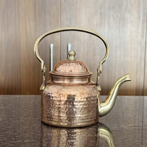 Könnte beinhalten: Eine Kupfer-Teekanne mit gehämmertem Muster, Messinggriff, Ausguss und Deckel. Die Kanne steht auf einer dunklen Oberfläche, die ihre Form widerspiegelt. Das Design ist vintage und dekorativ.