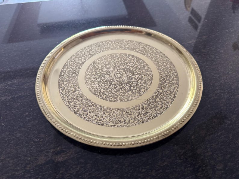 Puede incluir: Una bandeja redonda de metal dorado con un intrincado dise&ntilde;o floral en el centro. La bandeja tiene un borde con cuentas y un acabado pulido. Esta bandeja decorativa es adecuada para servir o exhibir.
