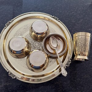 Puede incluir: Un juego de servir de metal dorado con una bandeja redonda, cuencos peque&ntilde;os, una cuchara y una taza. La bandeja y la taza tienen dise&ntilde;os decorativos. El juego est&aacute; dispuesto sobre una superficie oscura.