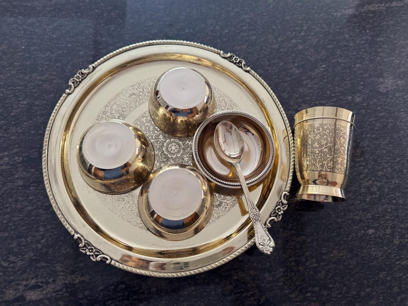 Puede incluir: Un juego de servir de metal dorado con una bandeja redonda, cuencos peque&ntilde;os, una cuchara y una taza. La bandeja y la taza tienen dise&ntilde;os decorativos. El juego est&aacute; dispuesto sobre una superficie oscura.