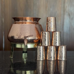 May include: A copper water dispenser with a hammered finish, a tap, and a brass stand. Six matching copper cups are stacked nearby. The copper items reflect the light, creating a warm aesthetic.