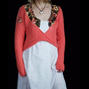 Könnte beinhalten: Ein korallenfarbener Strickcardigan mit floralem Besatz. Der Cardigan ist kurz geschnitten und hat einen V-Ausschnitt.