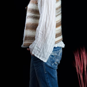May include: A woman wearing a brown and white striped knitted vest over a white blouse and blue jeans with rhinestone detailing.