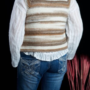 May include: A white and brown striped knitted vest with a ruffled white shirt underneath and blue jeans with embellished pockets.