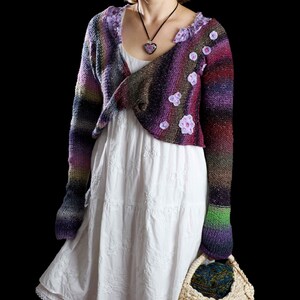 May include: A woman wearing a white dress with a colorful crocheted cardigan with purple, pink, and green accents. The cardigan has a heart-shaped pendant necklace and floral embellishments. She is holding a woven basket.
