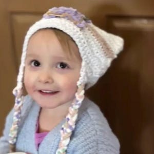May include: A child wearing a white crochet hat with lavender and tan accents and braided ties. The child is also wearing a light blue sweater and holding a container of cereal.