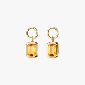 May include: A pair of gold earrings, each with a rectangular, yellow-orange gemstone. The earrings have a small, circular loop at the top and a rectangular gemstone set in a gold frame. The gemstones have a bright, translucent appearance.