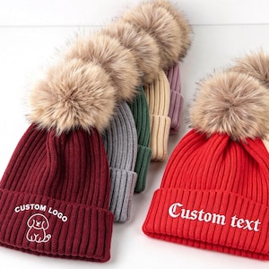May include: A collection of ribbed knit beanies in various colors including burgundy, gray, green, beige, red, and cream. Each beanie features a large faux fur pom-pom on top and a folded cuff. The burgundy beanie displays a dog graphic with the text CUSTOM LOGO, while the red beanie features the text Custom text.