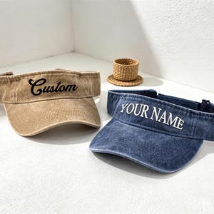May include: Two visors are shown, one in tan and one in blue. The tan visor has the word "Custom" embroidered in black. The blue visor has the words "YOUR NAME" embroidered in white. Both visors have a curved peak.