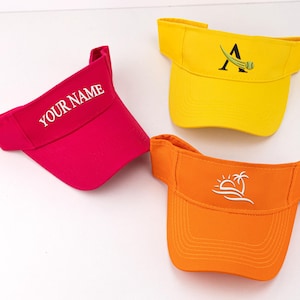 May include: Three visors in bright colors: fuchsia with "YOUR NAME" in white, yellow with a black "A" and tennis ball graphic, and orange with a white sun and palm tree design. These visors are ideal for sun protection.