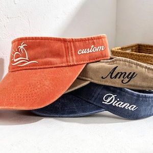 Peut inclure: Trois visières empilées, de couleurs orange, beige et bleu marine. La visière orange porte une broderie blanche représentant une scène de plage et le mot "custom". La visière beige porte "Amy", et la visière marine porte "Diana".