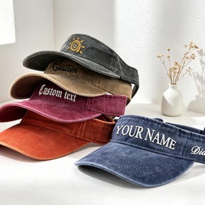 May include: Six visors in a stack, in shades of denim blue, burgundy, and brown. The visors have curved peaks and adjustable straps. Some have embroidered text, such as "YOUR NAME" and "Custom text", and a sun design.