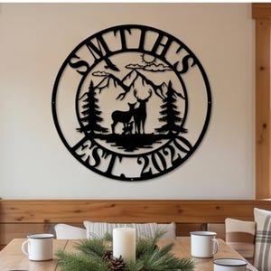 May include: A circular black metal wall sign featuring a laser-cut design of a deer family in a forest with mountains in the background. The text SMTH'S is arched at the top, and EST. 2020 is arched at the bottom of the circular frame.