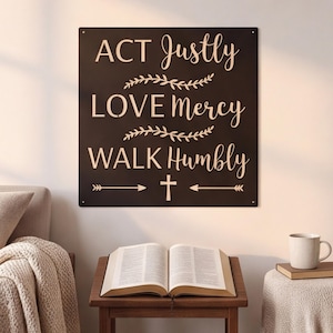 May include: A square black metal wall sign featuring the light-coloured text ACT Justly, LOVE Mercy, WALK Humbly. The design includes two horizontal leafy vine dividers, a small cross symbol, and an arrow graphic pointing in both directions.