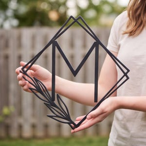 May include: A black metal wall sign featuring a large letter M centred within a diamond-shaped frame. The design includes a decorative leaf or branch element extending from the bottom left corner of the frame, creating a clean and geometric aesthetic.