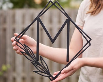 Può includere: Una decorazione da parete in metallo nero con una grande lettera M centrata all'interno di una cornice a forma di rombo. Il design include un elemento decorativo a forma di foglia o ramo che si estende dall'angolo inferiore sinistro.