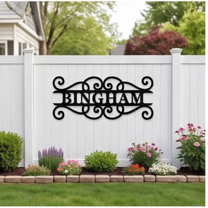以下が含まれることがあります： 黒い金属製の壁掛け看板。中央に大文字で「BINGHAM」と記され、左右対称のスクロール模様と渦巻き状の装飾が施されたフレームに収められている。白い縦板のフェンスに水平に取り付けられている。