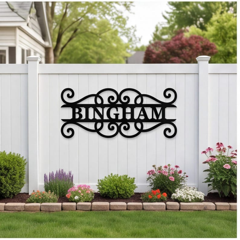 以下が含まれることがあります： 黒い金属製の壁掛け看板。中央に大文字で「BINGHAM」と記され、左右対称のスクロール模様と渦巻き状の装飾が施されたフレームに収められている。白い縦板のフェンスに水平に取り付けられている。