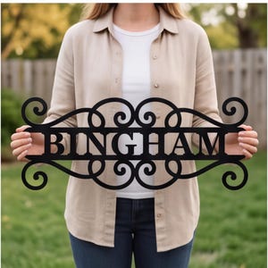 以下が含まれることがあります： 中央の長方形のフレームに大文字で「BINGHAM」と記された黒い金属製の看板。デザインには、中央のネームプレートの上下の端から伸びる装飾的なスクロール模様が施されています。