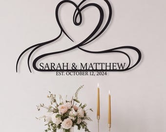 May include: A black metal wall sign featuring a continuous line art heart shape above the text SARAH & MATTHEW and EST. OCTOBER 12, 2024. The design is composed of smooth, flowing curves that form a stylised heart silhouette in a minimalist style.