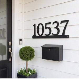 May include: A matte black metal house number sign displaying the digits 10537, featuring a horizontal base bar with mounting holes at each end, mounted on white horizontal siding above a black rectangular wall-mounted mailbox.