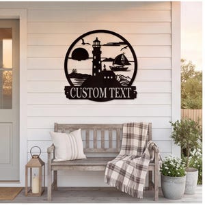 May include: A circular black metal wall sign featuring a lighthouse, a sailboat, and a sun on the horizon. Below the circular frame is a rectangular banner with the words CUSTOM TEXT in a serif font, designed for personalized lettering.