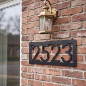 May include: A rectangular black metal house number sign displaying the digits 2523 in a bold sans-serif font. The sign is mounted on a red brick wall directly beneath a gold-toned outdoor wall lantern with a clear glass pane and a light bulb.