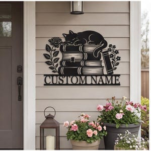 May include: A black metal wall sign featuring a sleeping cat resting on a stack of books, flanked by floral stems on both sides. Below the books, a horizontal bar displays the text CUSTOM NAME in a bold, capitalised serif font.