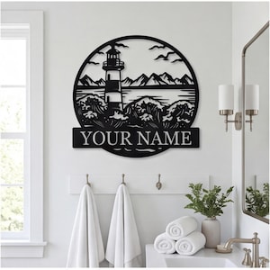 May include: A circular black metal wall art piece featuring a lighthouse, crashing waves, and mountain silhouettes. The design includes a rectangular banner at the bottom with the text YOUR NAME in a bold, sans-serif font, centered within the frame.