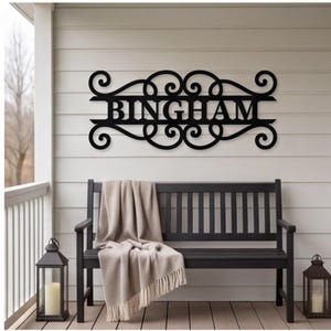 以下が含まれることがあります： 黒い金属製の壁掛け看板。中央に大文字で「BINGHAM」と記され、左右対称のスクロール模様と渦巻き模様で装飾された水平なフレームに収められており、明るい色の水平パネル壁に取り付けられています。