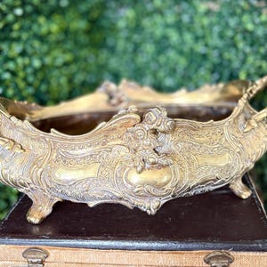 Puede incluir: Jardinera de metal dorado ornamentada con asas. La jardinera presenta diseños florales y de volutas detallados. Es rectangular con lados curvos y descansa sobre cuatro pequeños pies. El fondo es un seto verde.