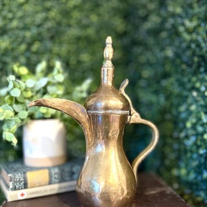 Puede incluir: Una cafetera de latón vintage con un pico largo y un asa curvada. La cafetera tiene un cuerpo bulboso y un adorno decorativo en la tapa. Se encuentra sobre una superficie oscura con un fondo verde borroso.