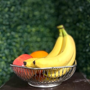 May include: A silver wire fruit basket filled with fresh fruit. The basket contains yellow bananas, a red apple, and an orange. The background is a blurred green hedge.