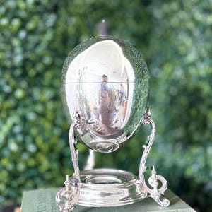 May include: A silver-plated egg-shaped serving dish with a lid, supported by an ornate stand. The dish reflects the surrounding environment. The stand features decorative scrollwork and a circular base, adding to its vintage aesthetic.