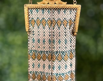 May include: An antique gold-toned metal mesh purse with a decorative frame and chain handle. The purse features a white background with a repeating pattern of teal and gold diamond shapes. The purse is suspended against a blurred green background.